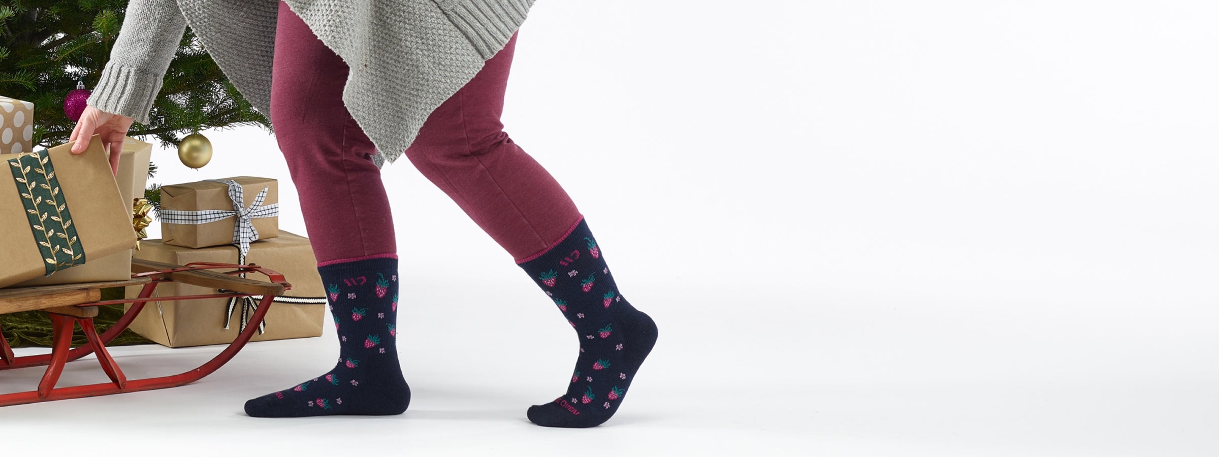 A person wearing Wide Open socks with strawberries on them, placing packages under a Christmas tree