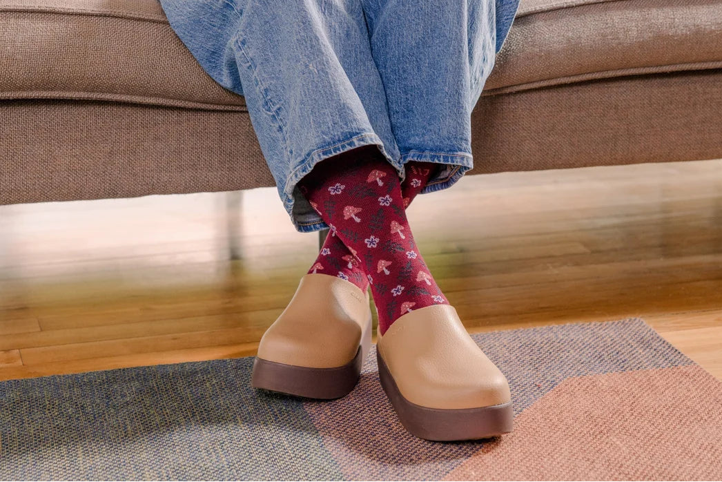 A person sitting on a brown couch wearing light blue jeans, burgundy patterned socks, and tan platform clogs on a rug over a wooden floor.