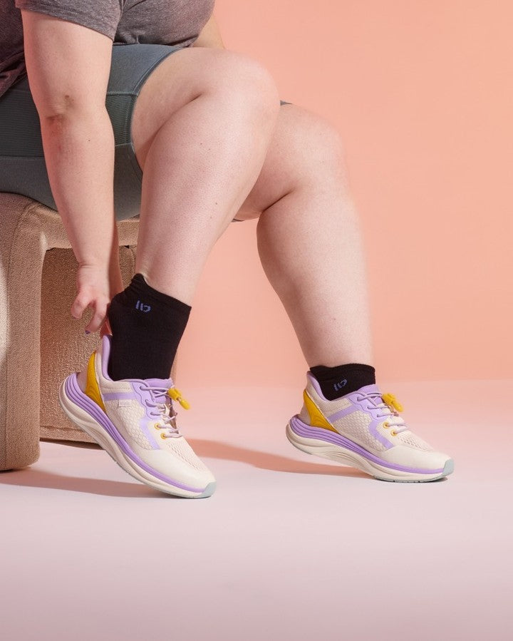 A person seated on a stool wearing a pair of athletic sneakers with cream, lavender, and yellow accents, paired with wide open black ankle socks, against a peach-pink studio background.